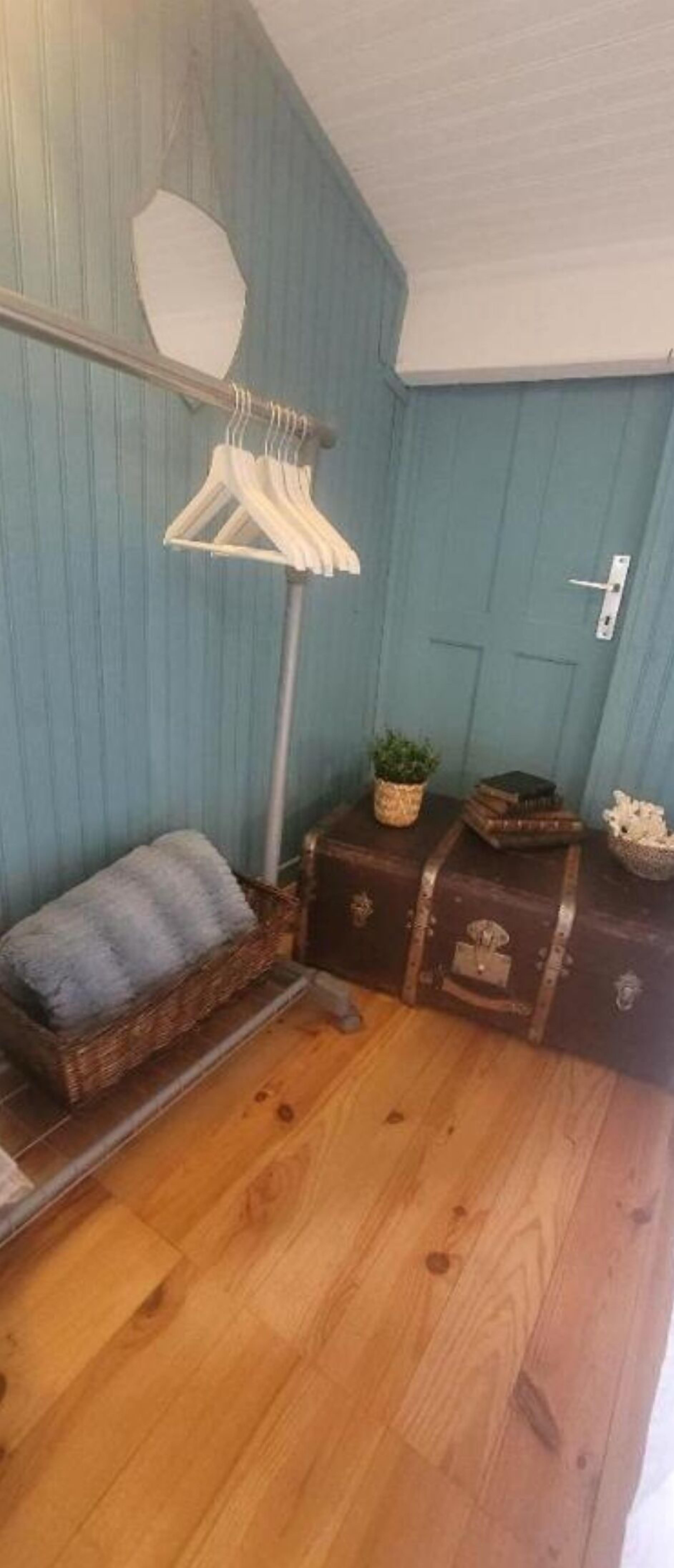 Ein Zimmer mit Holzboden, blauer Wand, weißer Decke, einem Bett mit blauer Decke, einer hölzernen Bank, einem kleinen Tisch mit Pflanze und einer Tür.