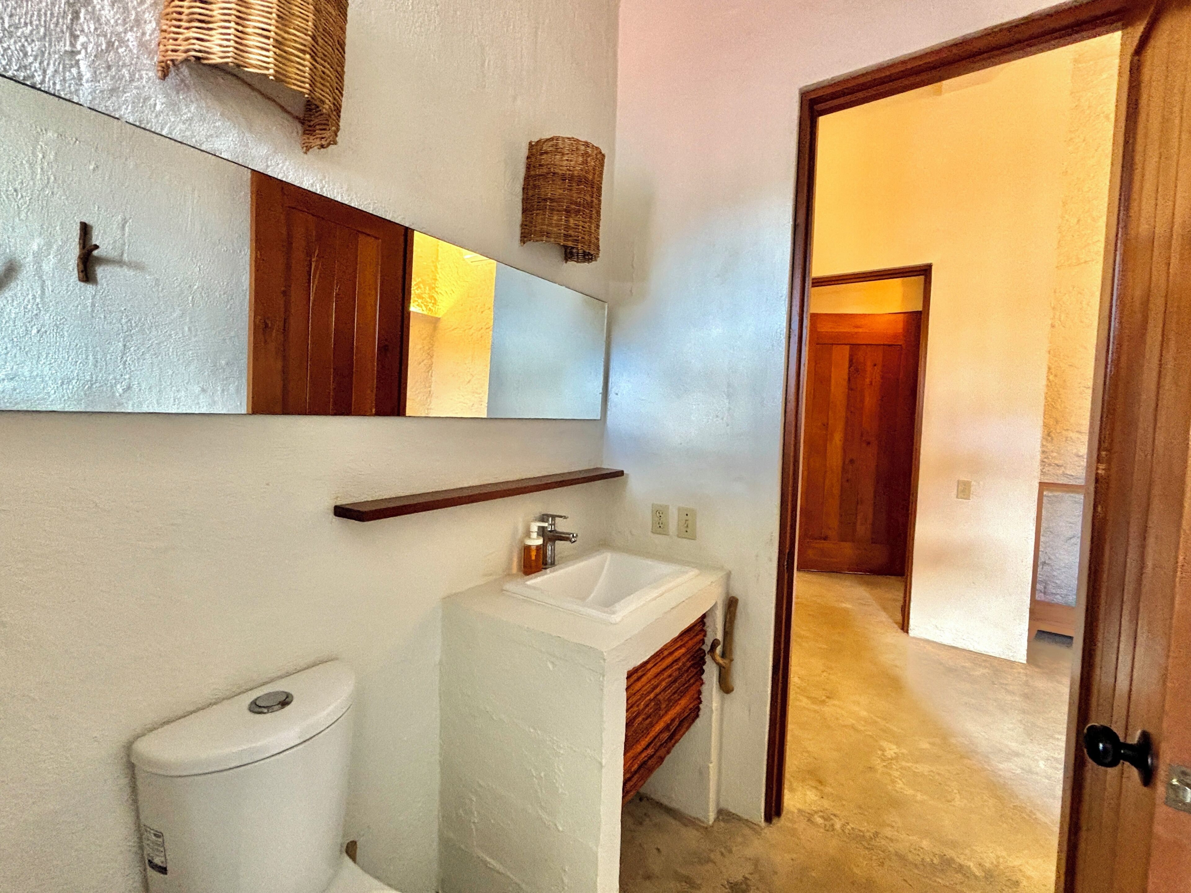 A bathroom with a white toilet, a sink, and a mirror. There is a wooden door leading to another room.