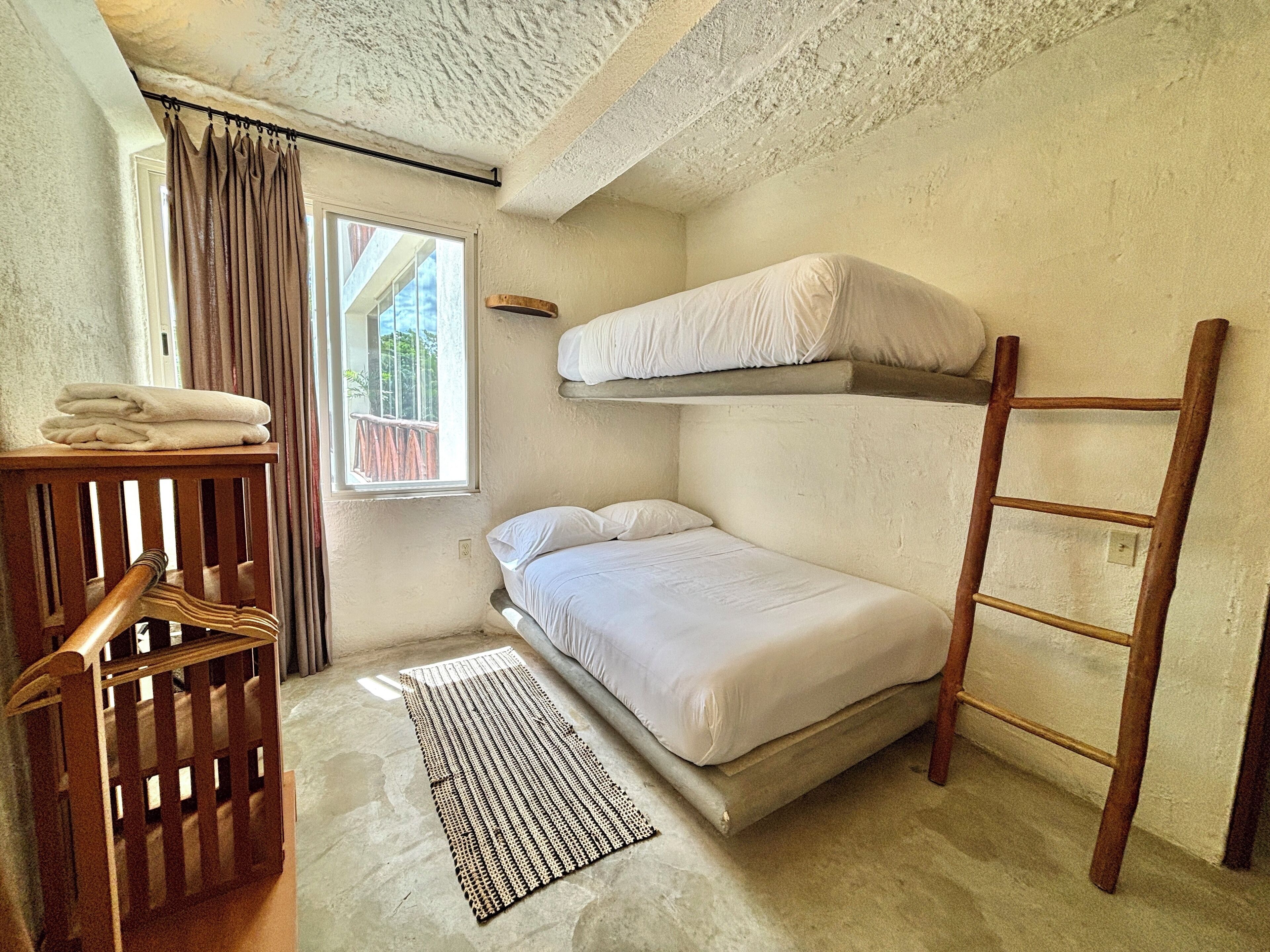 A compact room with a bunk bed, a wooden ladder, a small table with towels, and a window with curtains.