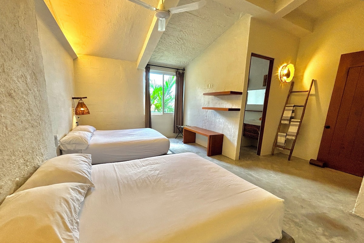 A bedroom with two beds, a ceiling fan, a bench, a ladder, and a window with curtains.