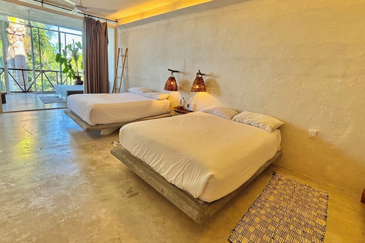 A modern hotel room with two beds, a large window with a view of greenery, and a ceiling fan.