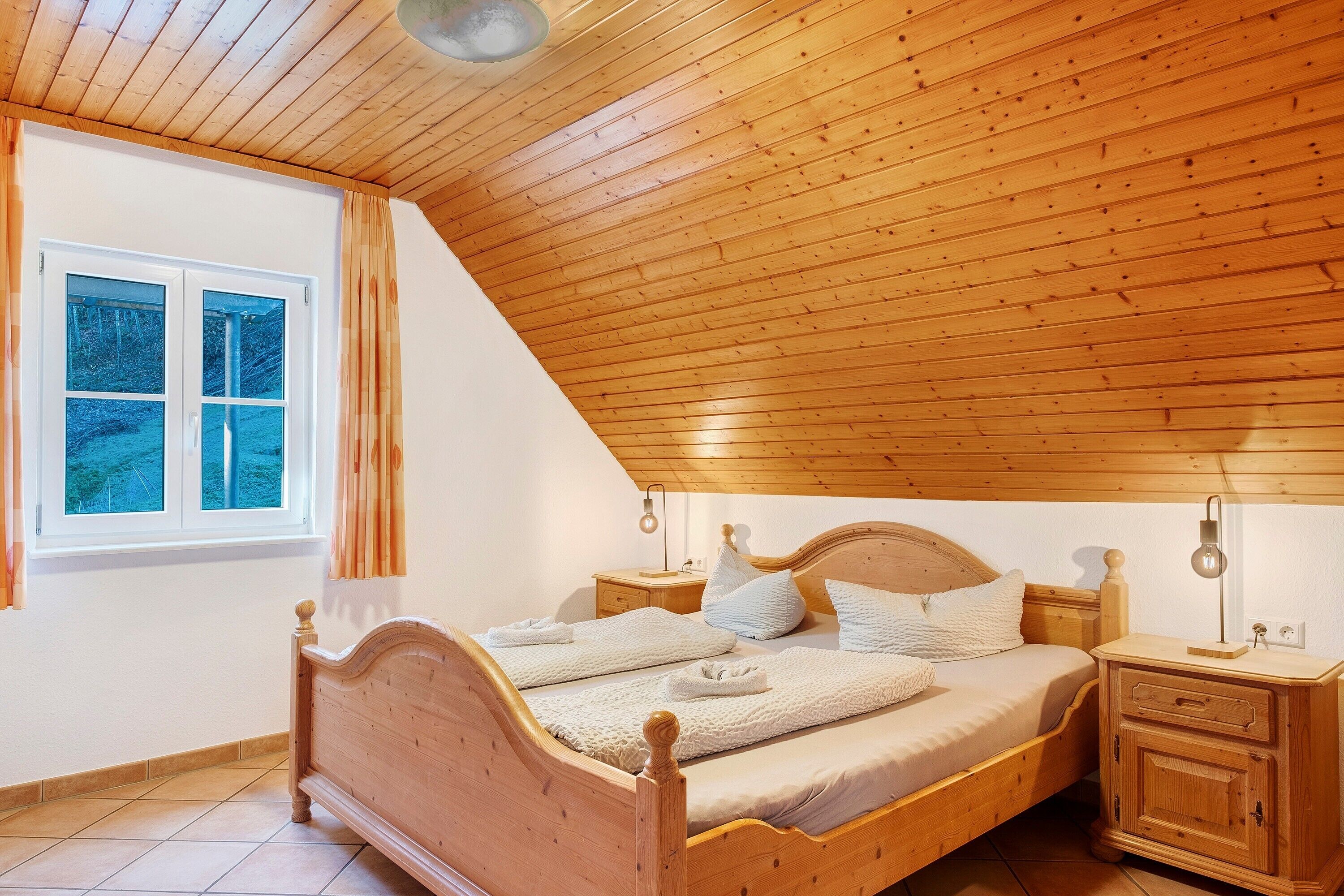 Un dormitorio con techo de madera inclinado, una cama de madera con cabecera, una mesita de noche con lámpara y una ventana con cortinas.