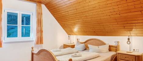 Un dormitorio con techo de madera inclinado, una cama de madera con cabecera, una mesita de noche con lámpara y una ventana con cortinas.