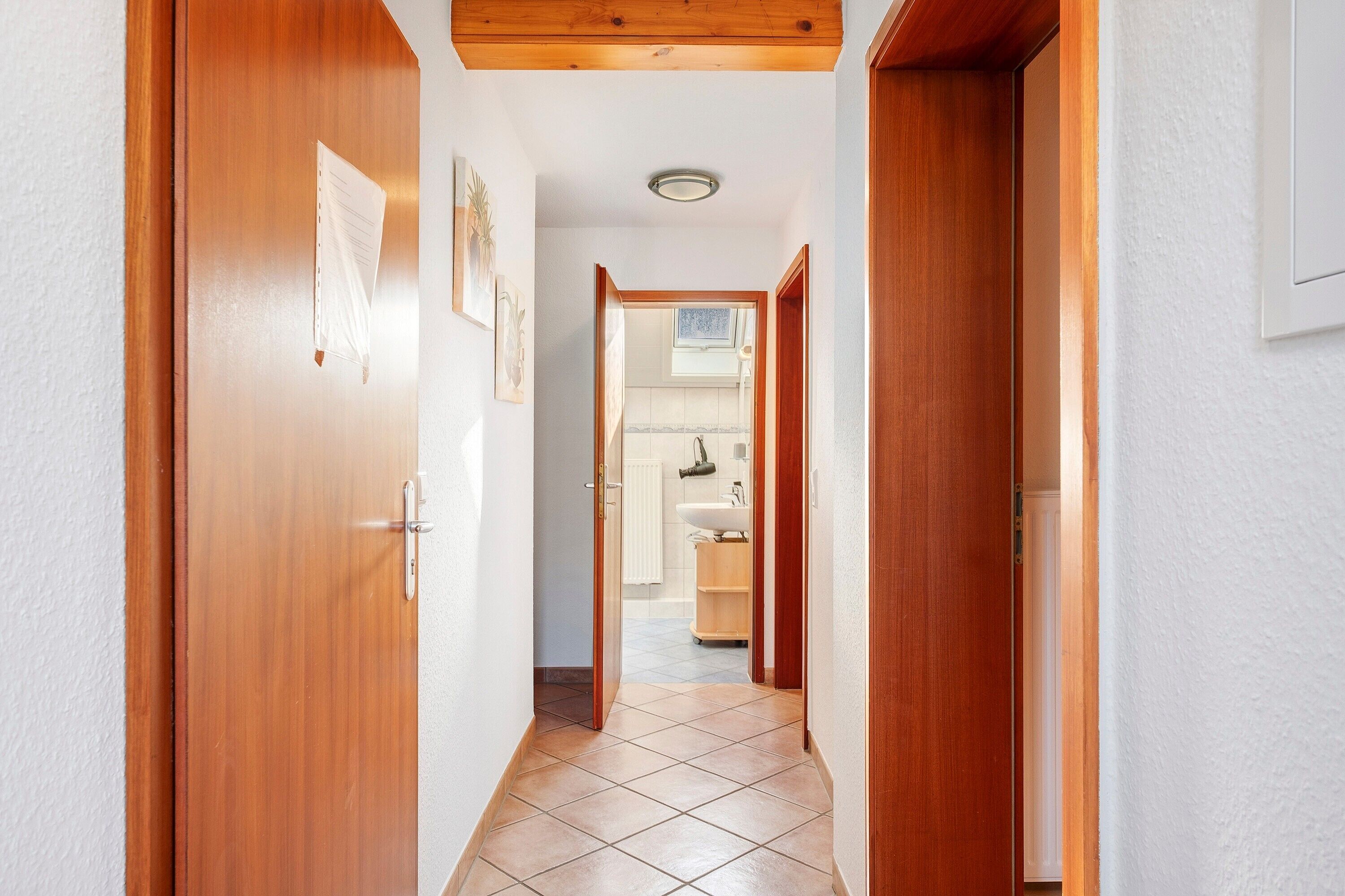 Ein Korridor mit Holztüren, Fliesenboden und Blick in ein Badezimmer mit Waschbecken und Spiegel.