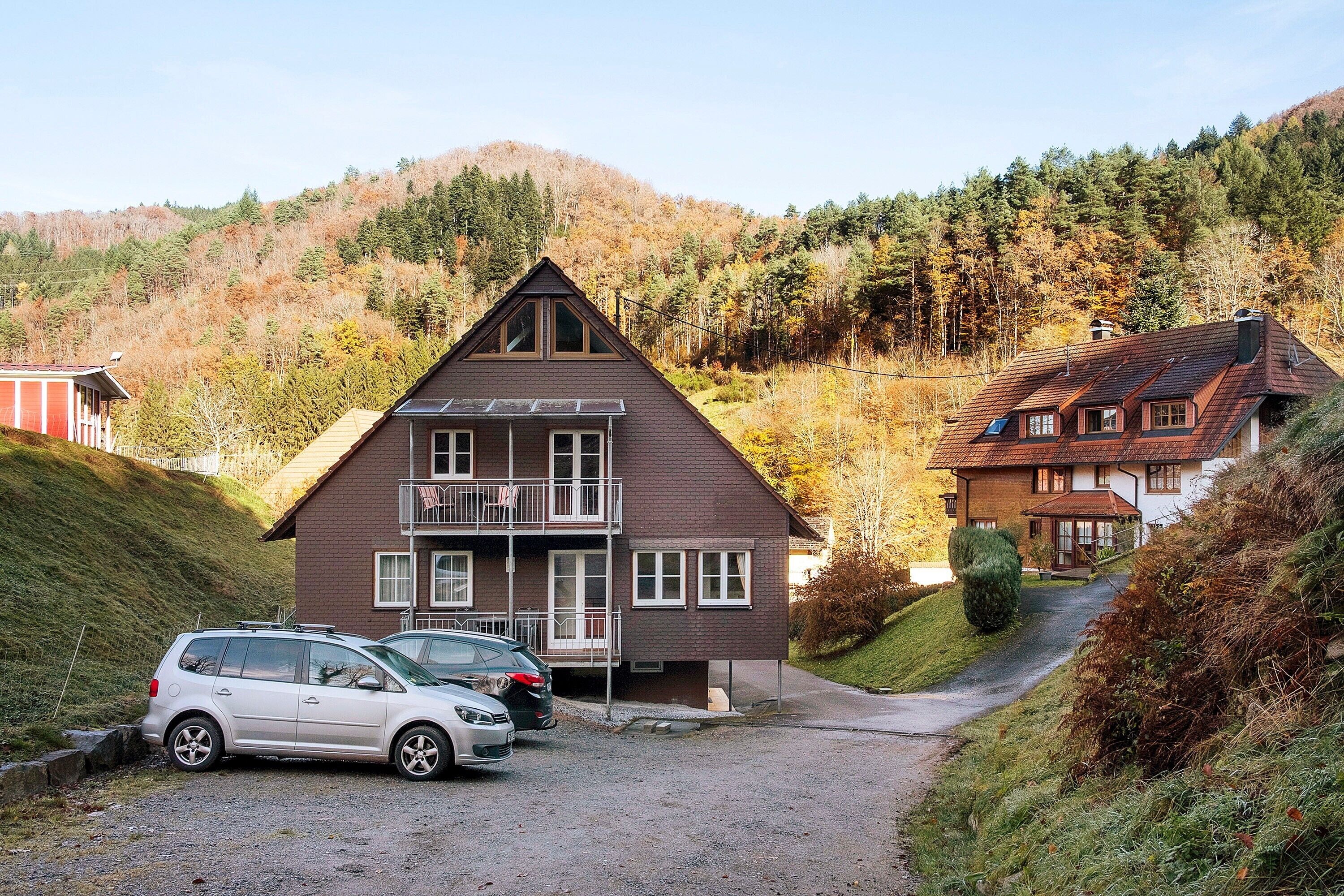 A two-story house with a sloped roof, a balcony, and a driveway leading to parked cars.