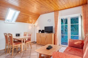 A cozy room with a wooden ceiling, a dining table with chairs, a sofa, a TV on a cabinet, and a window with curtains.