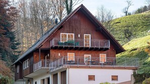 A two-story wooden building with a balcony, a covered entrance, and a hillside backdrop. - Apartment 'Vogelnest' with Mountain View, Shared Pool and Wi-Fi – Not Suitable for Dogs (Wolfach)