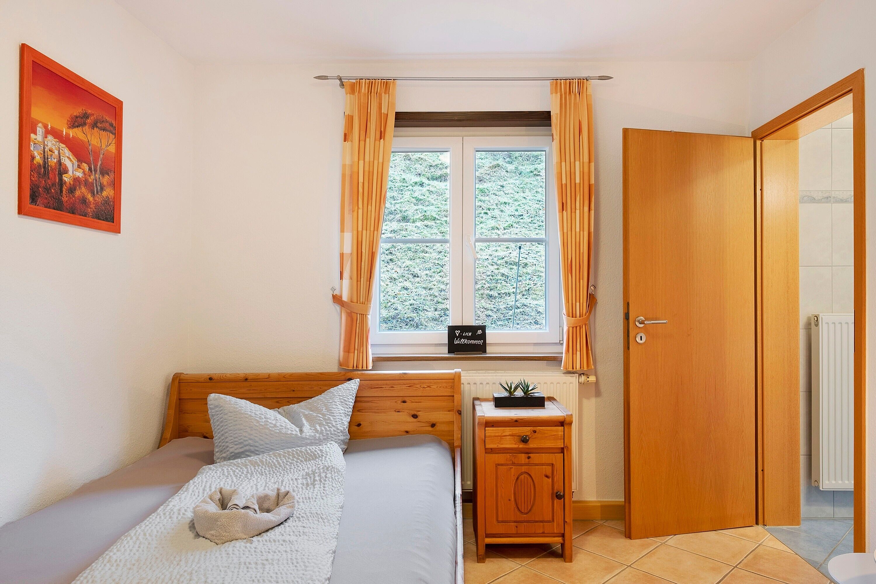 A bedroom with a bed, a bedside table, a nightstand, a window with curtains, a door, and a radiator.