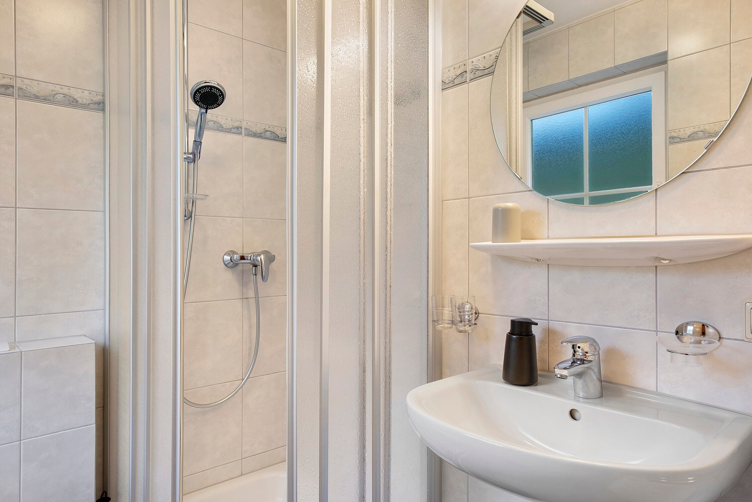 Een badkamer met een douche, wastafel, spiegel en een plank met toiletartikelen.