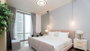 A modern hotel room with a large bed, a bedside table with a lamp, a small seating area, and a view of the city through large windows. - 2BR Waterfront Apartment  Nr Downtown (Business Bay)