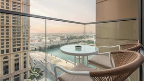 A balcony with a glass railing, a round table, and two wicker chairs. A view of a cityscape with buildings and a river is visible through the railing. - 2BR Waterfront Apartment  Nr Downtown (Business Bay)