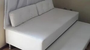 Un lit blanc avec une tête de lit et deux oreillers, dans une pièce aux murs unis et dotée d’une fenêtre avec des rideaux.