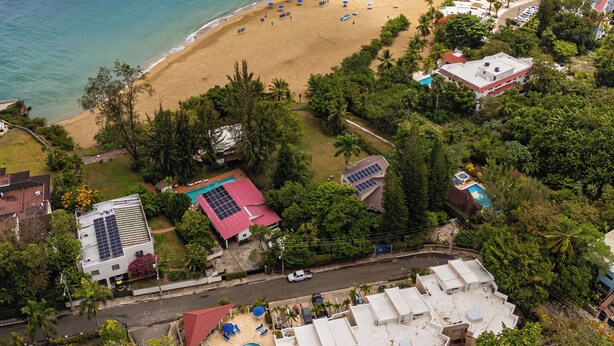 Luftaufnahme eines Küstenresorts mit Strand, Schwimmbad und mehreren Gebäuden mit Solaranlagen.