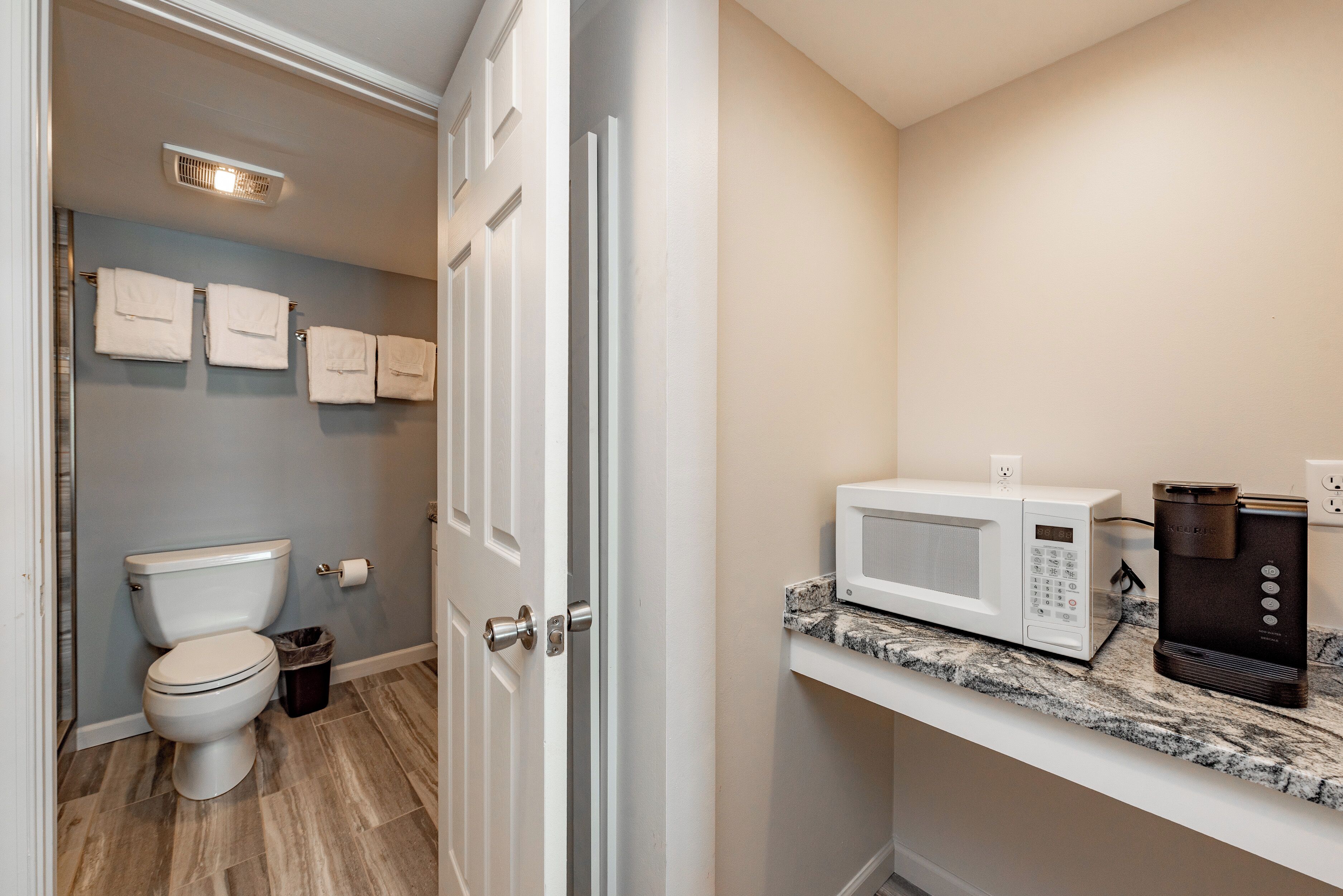 A bathroom with a toilet, a trash can, a towel rack with towels, a microwave, and a coffee maker on a countertop.