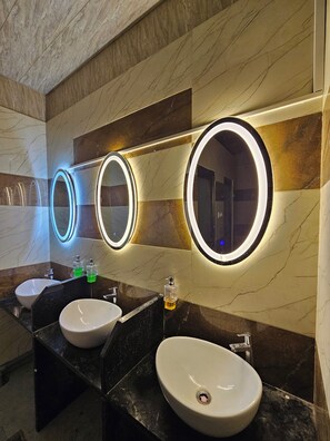 A modern bathroom with three sinks, each with a mirror above. The walls are adorned with marble tiles in various shades. - Kashi Vandanam (Varanasi)