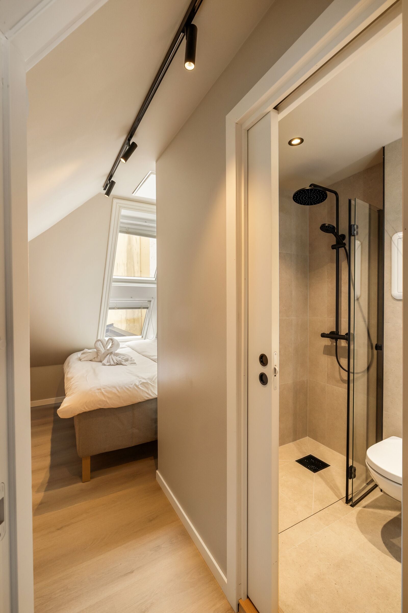 A modern bathroom with a glass shower enclosure, a toilet, and a small window above a bed.