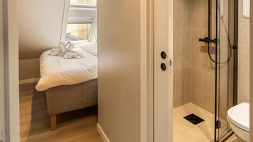 A modern bathroom with a glass shower enclosure, a toilet, and a small window above a bed.