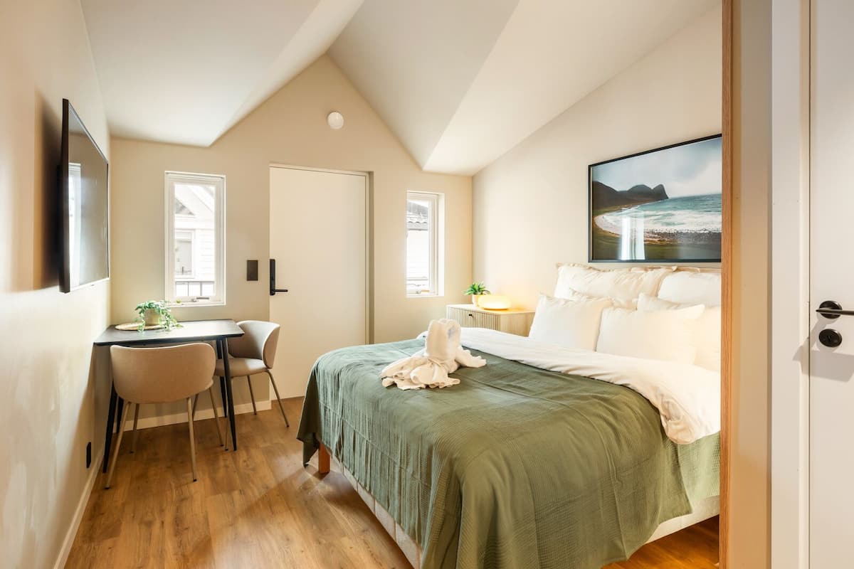 Un dormitorio con cama, escritorio y silla, ventana con vistas, y un cuadro en la pared.