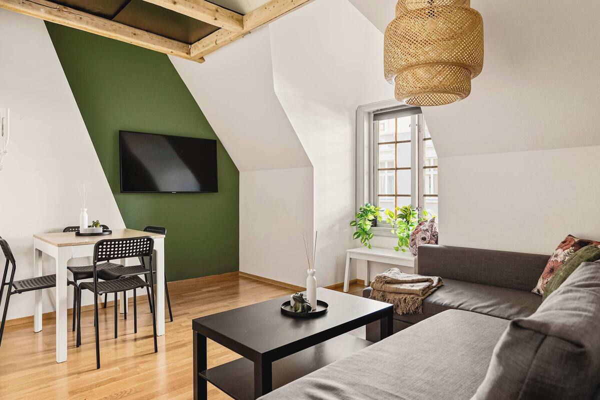 Una sala de estar moderna con comedor, un televisor montado en una pared verde, un ventanal grande y una lámpara colgante.