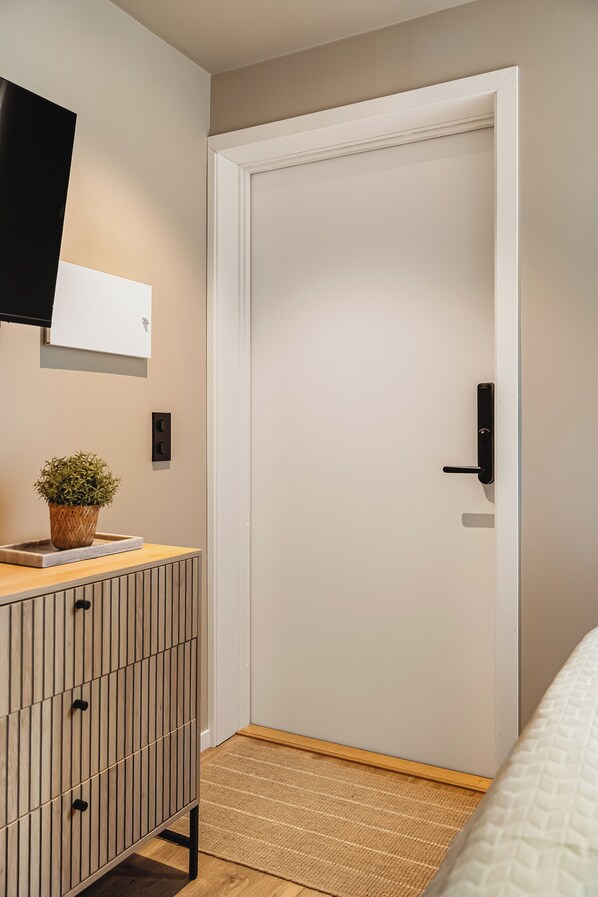 A modern bedroom with a wooden dresser, a potted plant, a white door, and a bed with a patterned bedspread. - Sigurds gate 8 (Bergen)