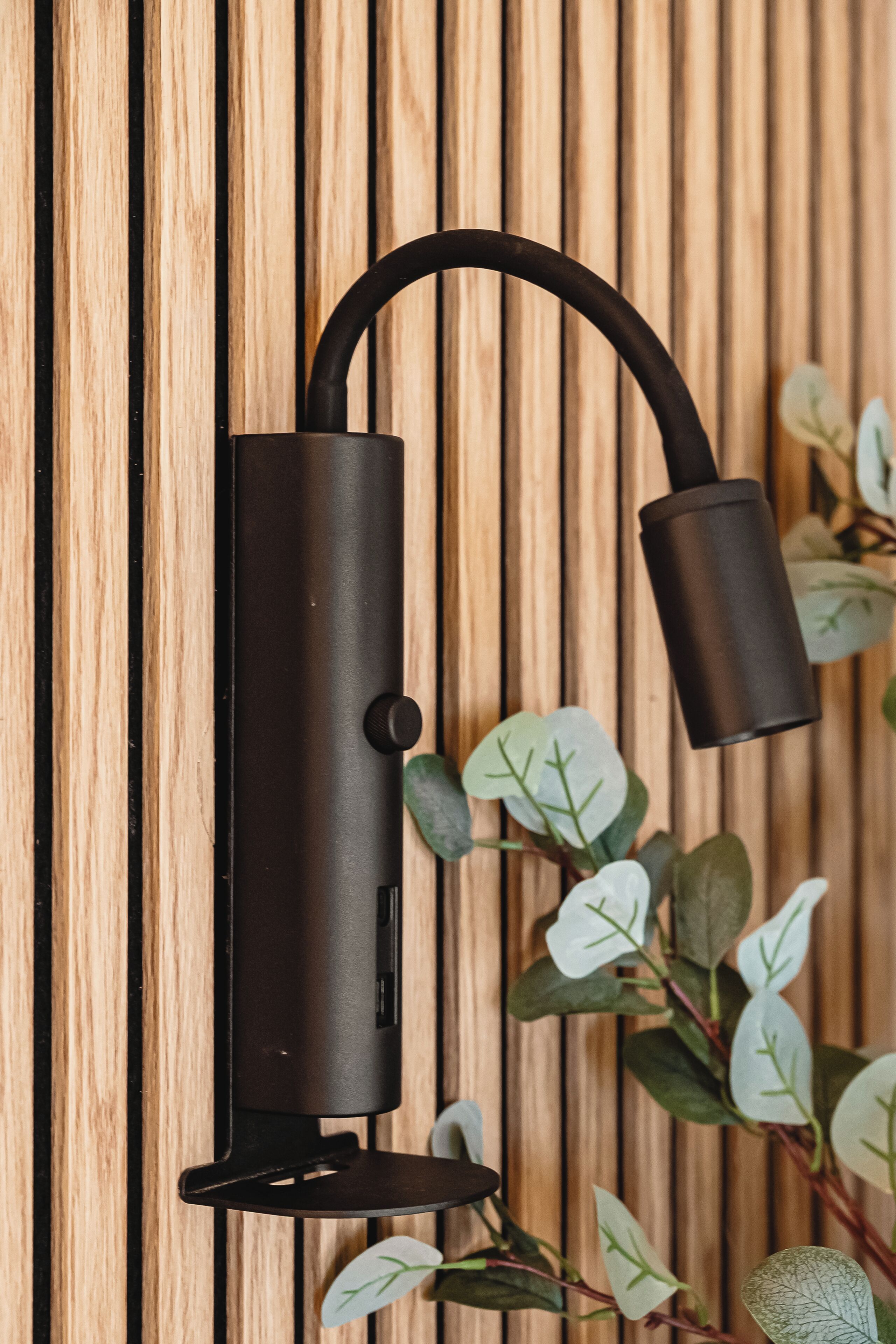 A wall-mounted light fixture with a curved arm and a base, set against a wooden panel background.