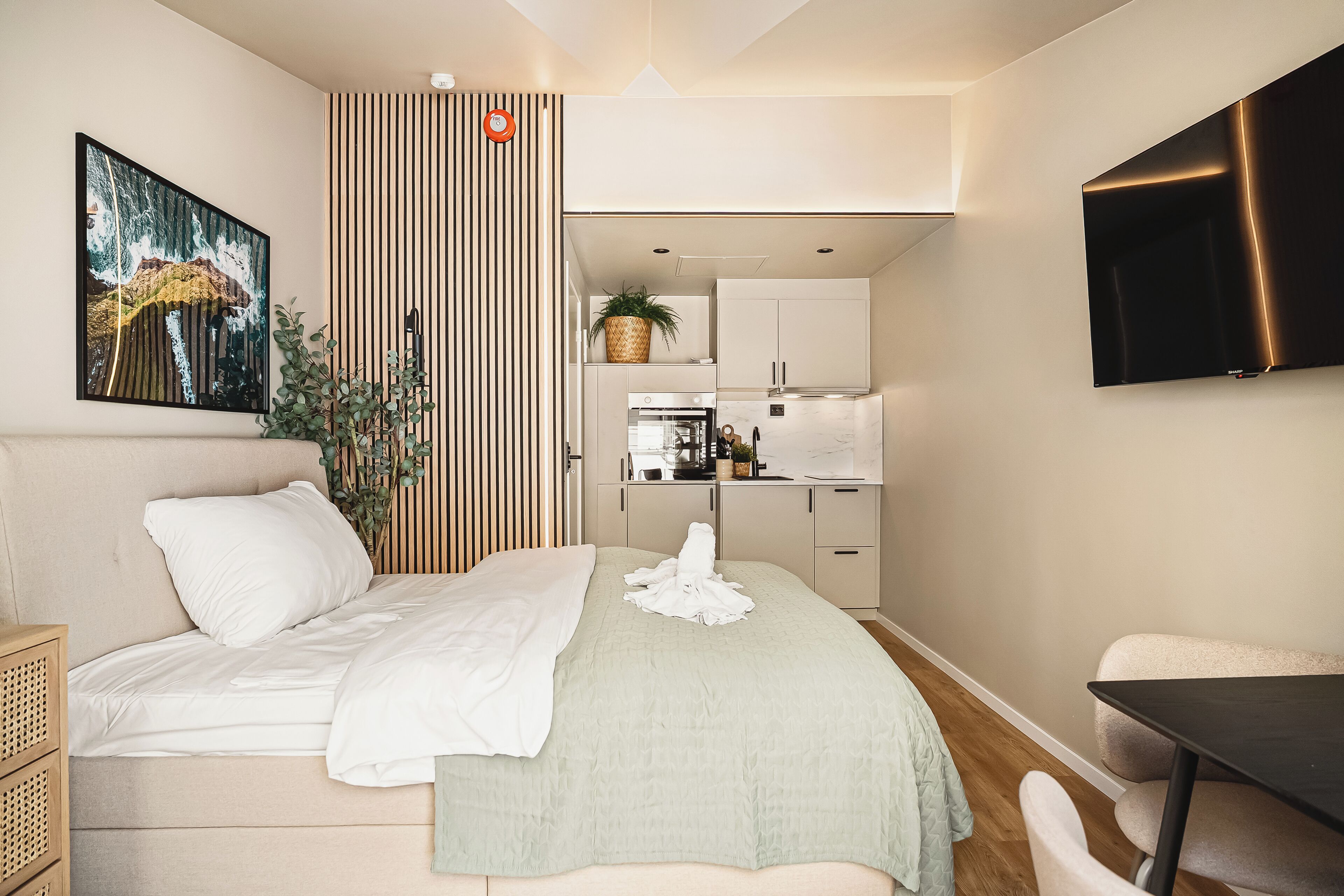 Moderni hotellihuone, jossa on sänky, työpöytä, tuoli, televisio ja pieni keittiö.