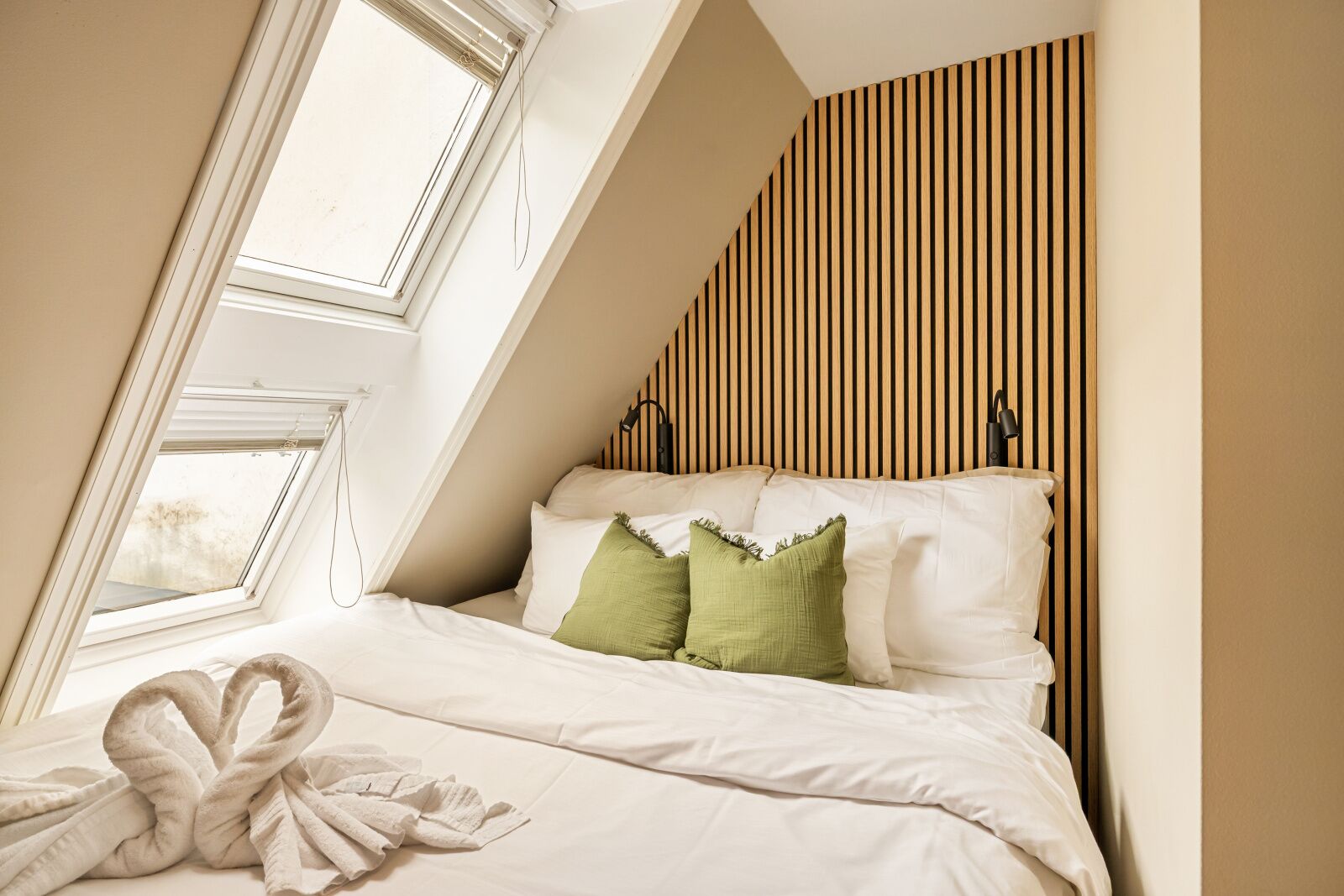 Un dormitorio con techo inclinado, una cama con ropa de cama blanca y almohadas verdes, una toalla sobre la cama y un panel de pared de madera.