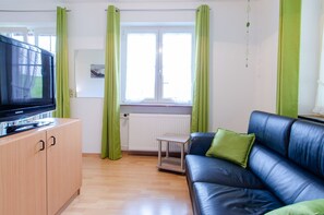 Une pièce avec un téléviseur posé sur un meuble en bois, un canapé en cuir bleu agrémenté de coussins verts, une petite table et une fenêtre ornée de rideaux verts.