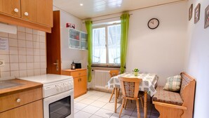 Una cucina con piano cottura, forno e mobili in legno. Si nota un tavolo da pranzo con sedie e una panca. È visibile una finestra con tende.