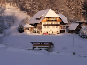 Un grand immeuble à plusieurs étages, dont le toit est recouvert de neige, entouré d’un paysage enneigé et d’arbres.