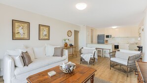 A modern living room with a white sofa, a wooden coffee table, and a dining area with wicker chairs. There is a kitchen in the background with white cabinets and stainless steel appliances.