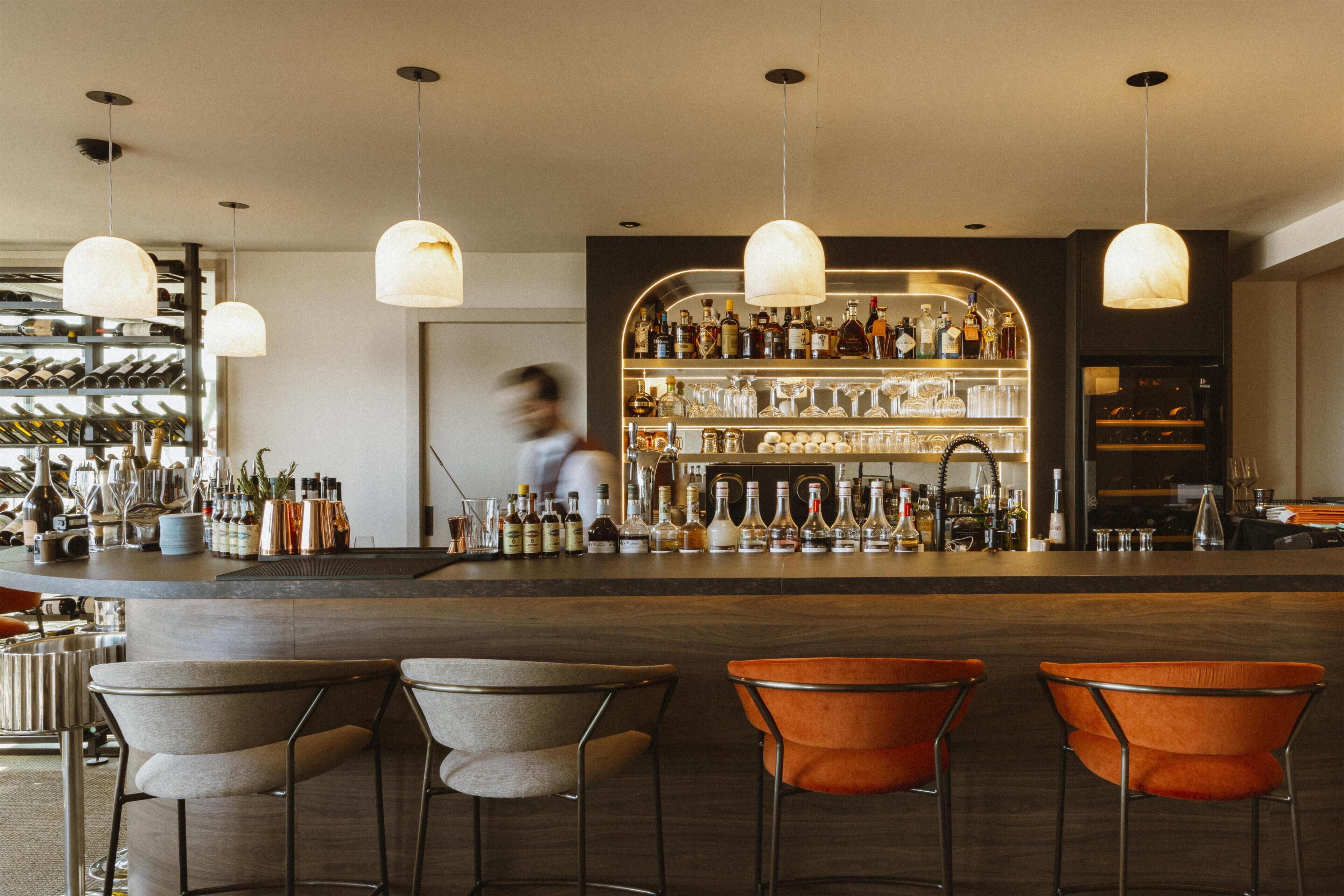 Eine Bar mit Tresen, zahlreichen Flaschen, Gläsern und drei Barhocker.