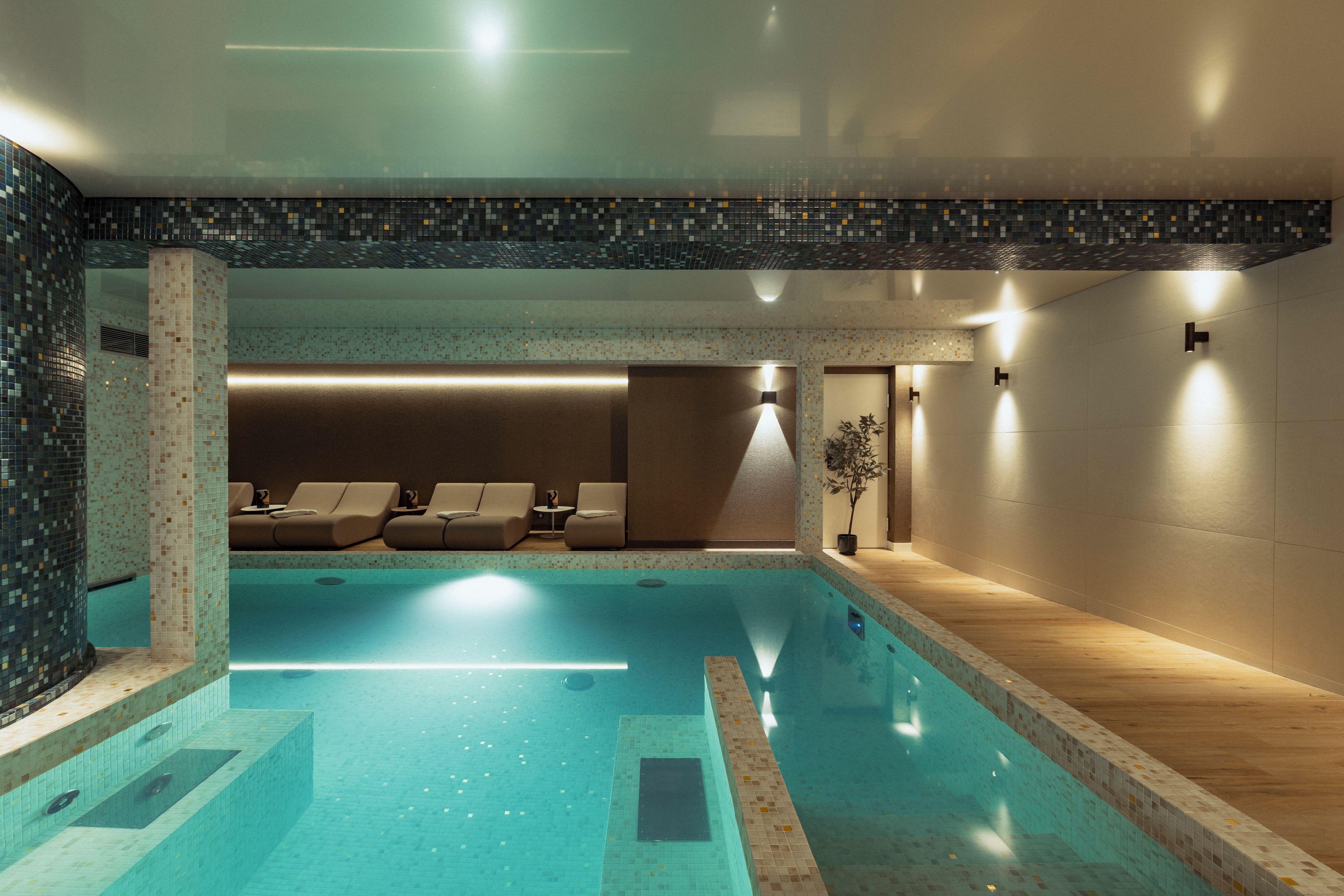 Une piscine intérieure moderne, avec un sol et des murs carrelés, un espace détente avec des sièges et une plante en pot.