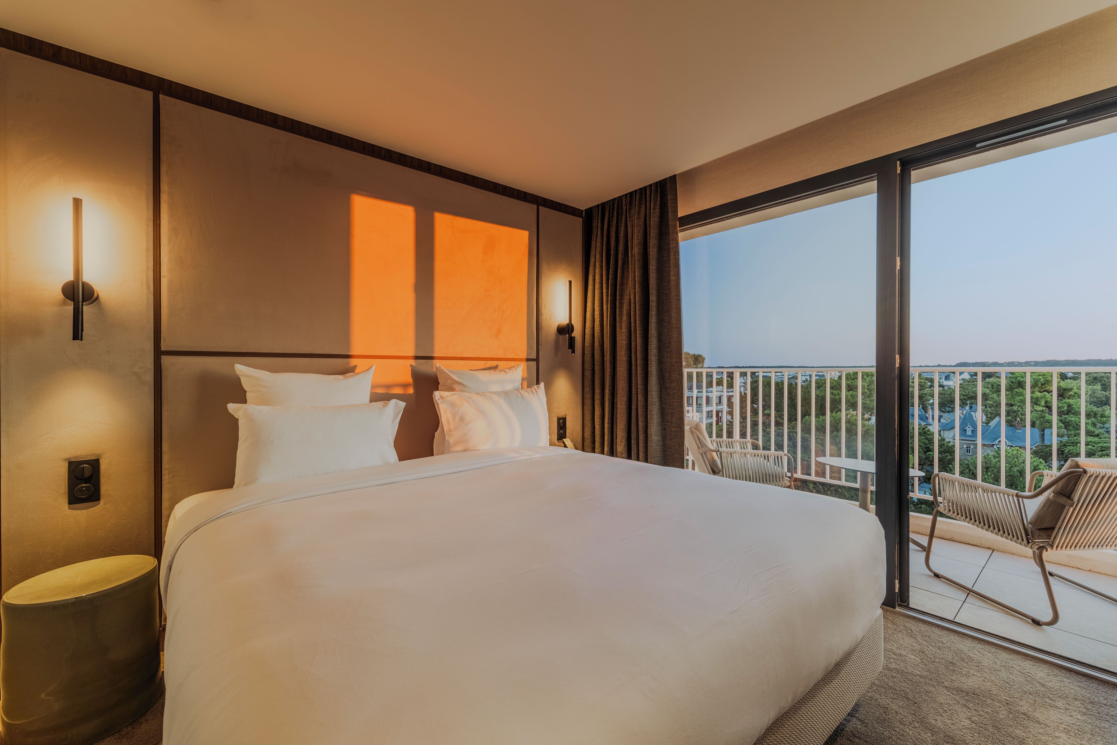 Habitación de hotel con una cama grande, vistas a la ciudad y un balcón con sillas.