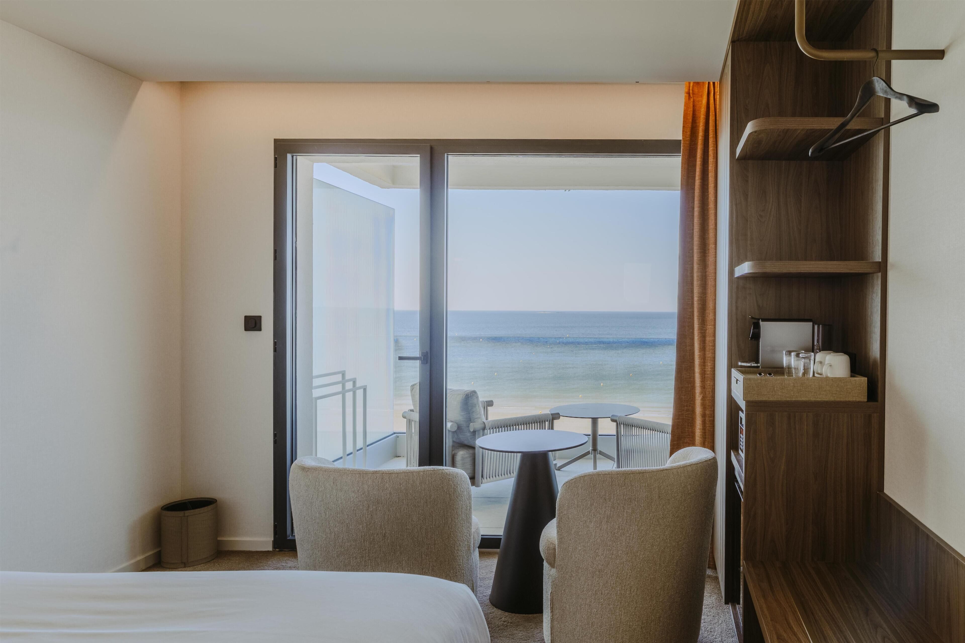 A hotel room with a bed, two chairs, a small table, a wooden shelf with items, and a view of the sea through a glass door.
