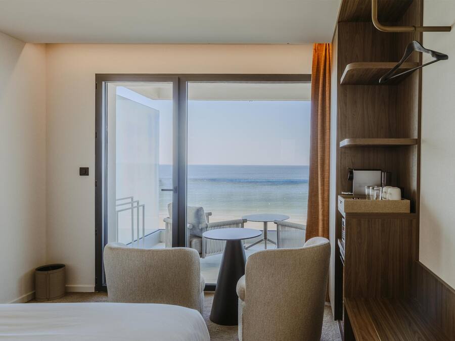 A hotel room with a bed, two chairs, a small table, a wooden shelf with items, and a view of the sea through a glass door.