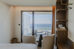 A hotel room with a bed, two chairs, a small table, a wooden shelf with items, and a view of the sea through a glass door. - Hotel & Spa Amoria La Baule, A Member Of Radisson Individuals (La Baule-Escoublac)