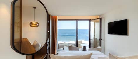Habitación de hotel con cama, televisor, vista al mar y balcón con mobiliario exterior.