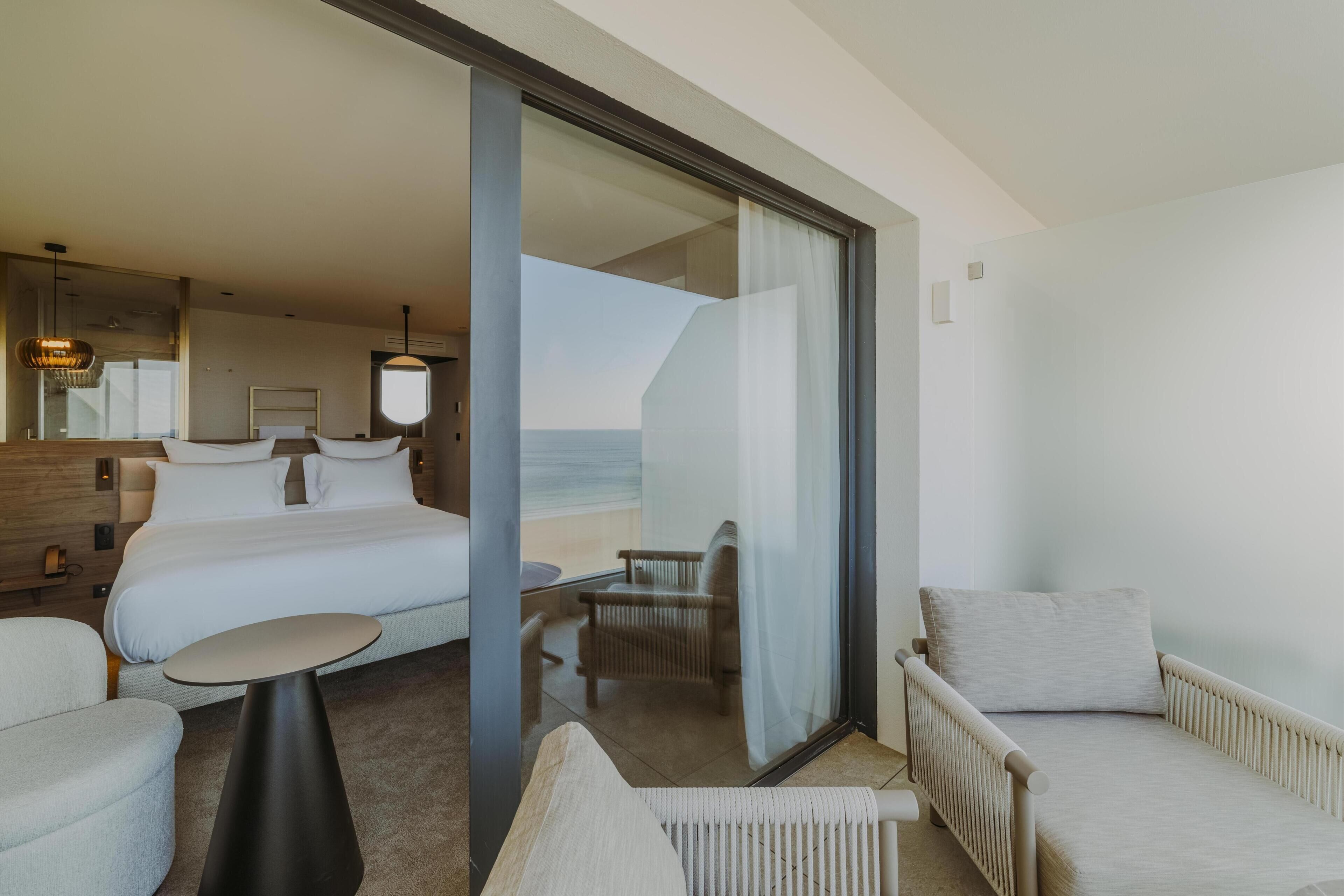 Une chambre d’hôtel avec un lit, une table ronde, une chaise et une vue sur la mer, visible à travers une porte vitrée.