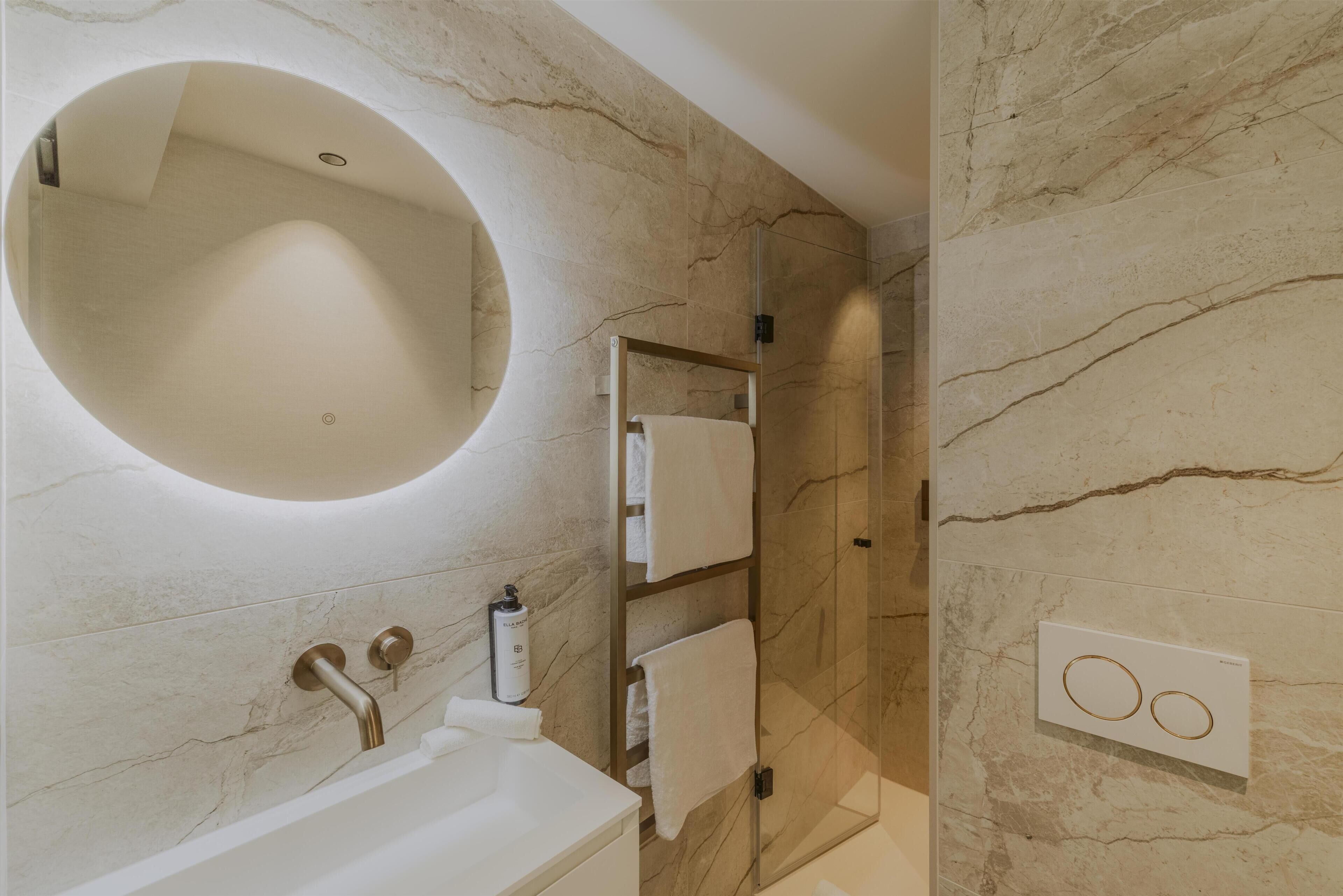 Une salle de bain avec un miroir rond, un lavabo blanc, un porte-serviettes et une cabine de douche en verre.