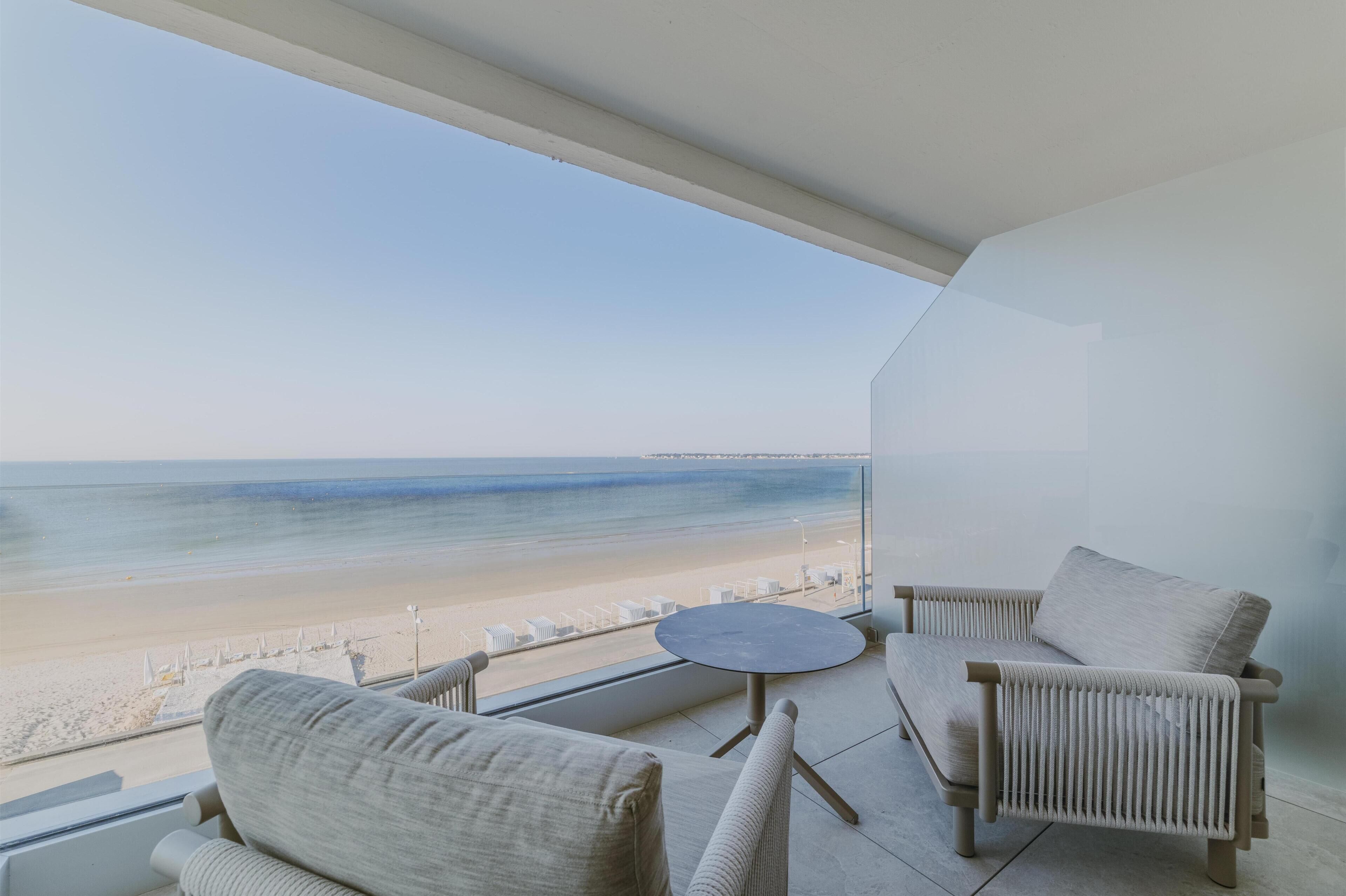 Un balcon donnant sur la plage, deux chaises, une petite table ronde et un mur blanc.