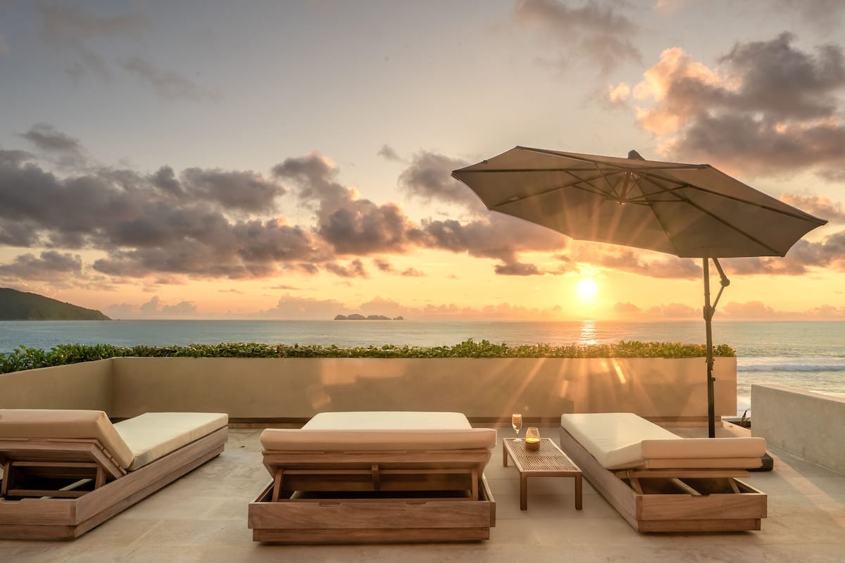 A beachside lounge with a sunset view, including lounge chairs, a table with a candle, and an umbrella.