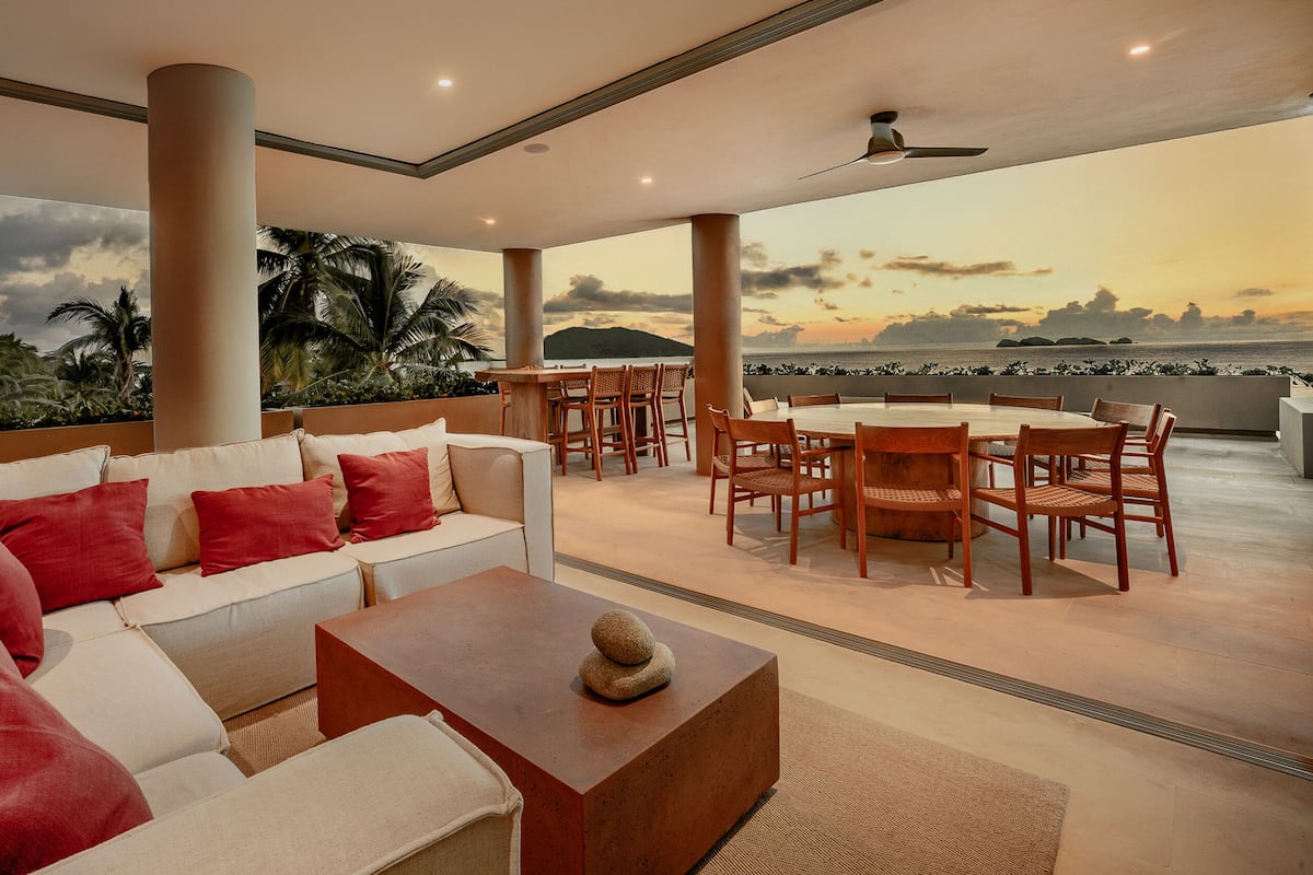 A modern living room with a sofa, coffee table, and dining area with chairs. There is a view of the ocean and palm trees through large windows.