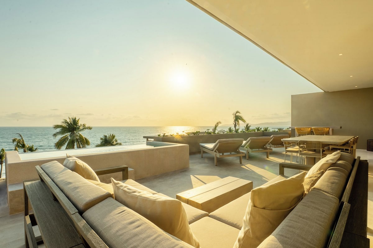 A modern outdoor lounge area with a view of the ocean, featuring a sofa set, chairs, and a table.