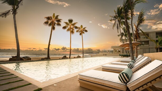 Zona junto a la piscina con sillones de descanso y vista al océano al atardecer.