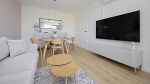 A modern living room with a white sofa, a dining table with chairs, a TV mounted on the wall, and a radiator.