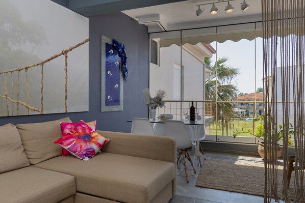 A modern living room with a beige sofa, a glass dining table, and a view of a residential area through large windows. - Casa Evita by Rentals Pro - Toroni Halkidiki (Sithonia)
