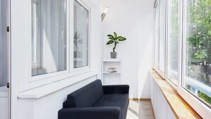 Una habitación estrecha con un sofá negro, un estante blanco pequeño con una planta y una ventana que da a vistas de vegetación.