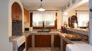 A kitchen with a stove, microwave, sink, and a window with curtains. - Azalia House by Rentalspro (Nea Propontida)