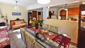 A dining area with a glass table, chairs, and a red table runner. There is a sofa with cushions and a bookshelf in the background. - Azalia House by Rentalspro (Nea Propontida)